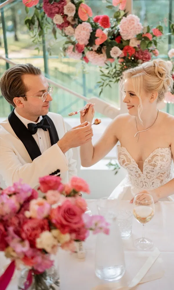 Bräutigam und Braut genießen gemeinsam ihr Hochzeitsmenü vor einer Kulisse aus rosa und pinken Blumen.