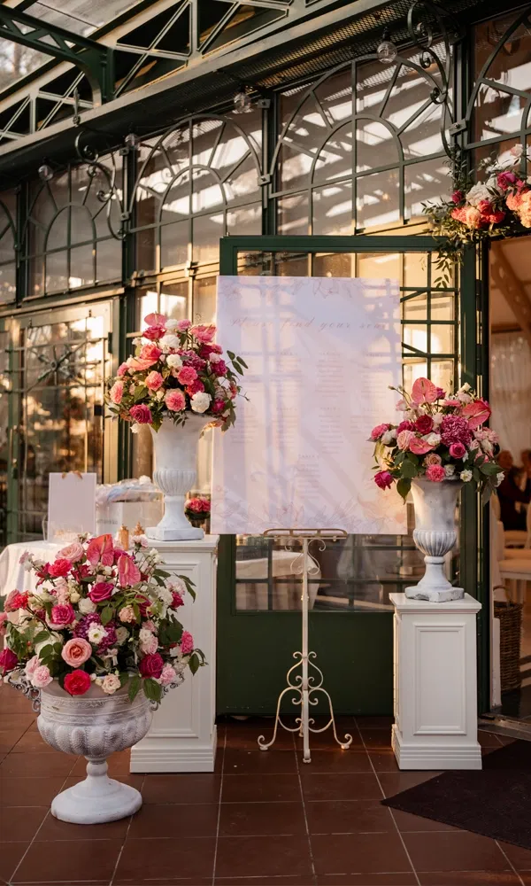 Festlich dekorierter Eingang mit üppigen Blumenarrangements in Rosa- und Pinktönen und einem Sitzplan auf Staffelei.