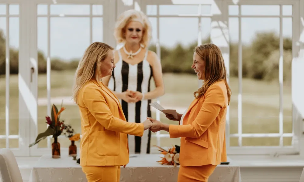 Hochzeitspaar in leuchtend orangefarbenen Anzügen tauscht bei der Zeremonie Blicke und Berührungen, im Hintergrund die Traurednerin.