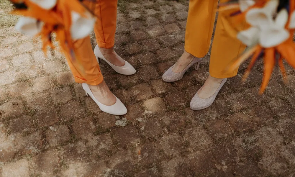 Detailaufnahme von zwei Paar Pumps in Beige und Glitzeroptik, getragen zu leuchtend orangefarbenen Hosen.