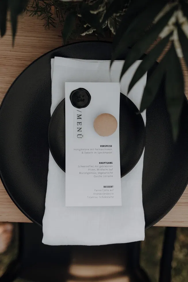 Stilvolles Hochzeitsmenü auf schwarzem Teller mit Leinenserviette, Siegel und minimalistisch-eleganter Tischdekoration