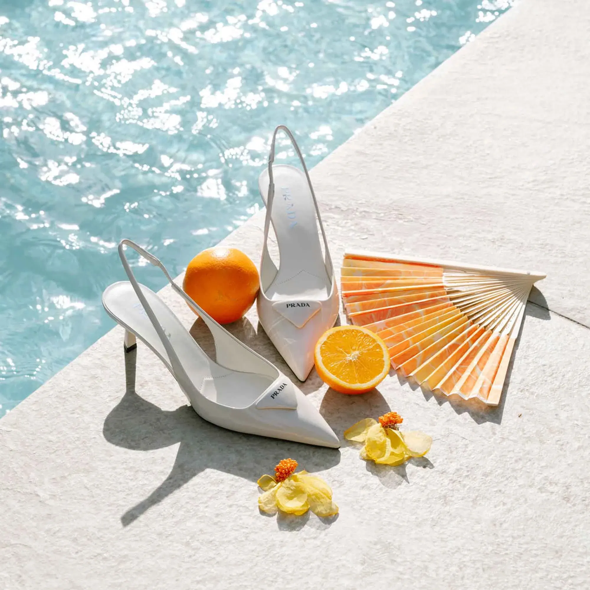 Weiße Prada-High-Heels am Pool, dekoriert mit einer Orange, einem Fächer und gelben Blüten für sommerliche Hochzeitsinspiration.
