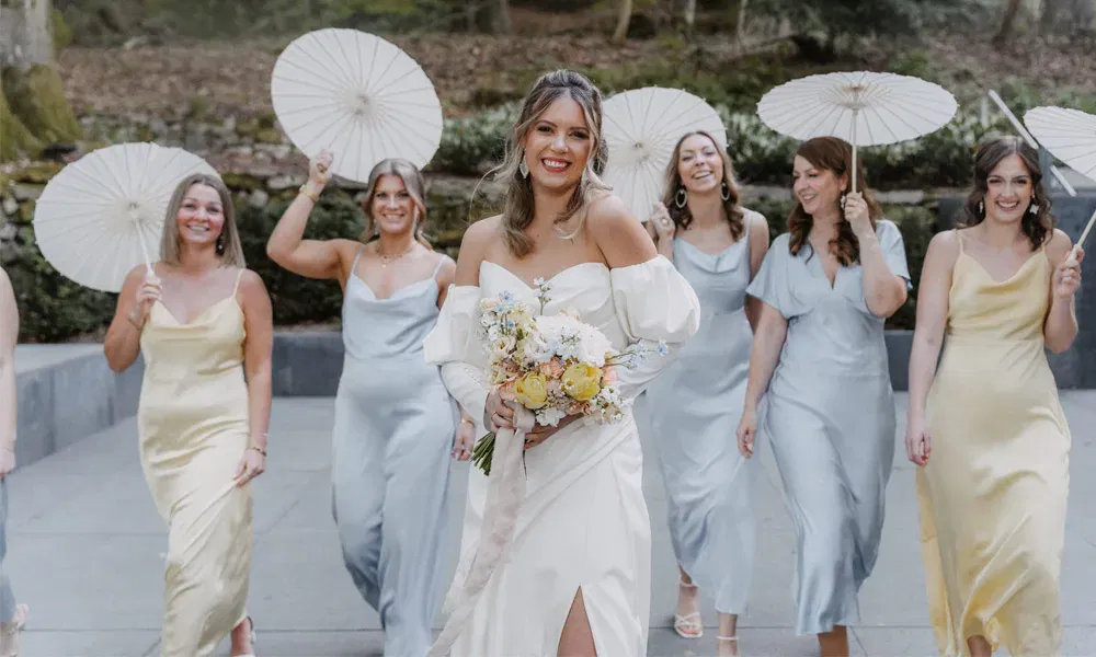 Braut in elegantem weißen Kleid mit Blumenstrauß, umgeben von Brautjungfern in hellblauen und gelben Kleidern, die weiße Sonnenschirme tragen.
