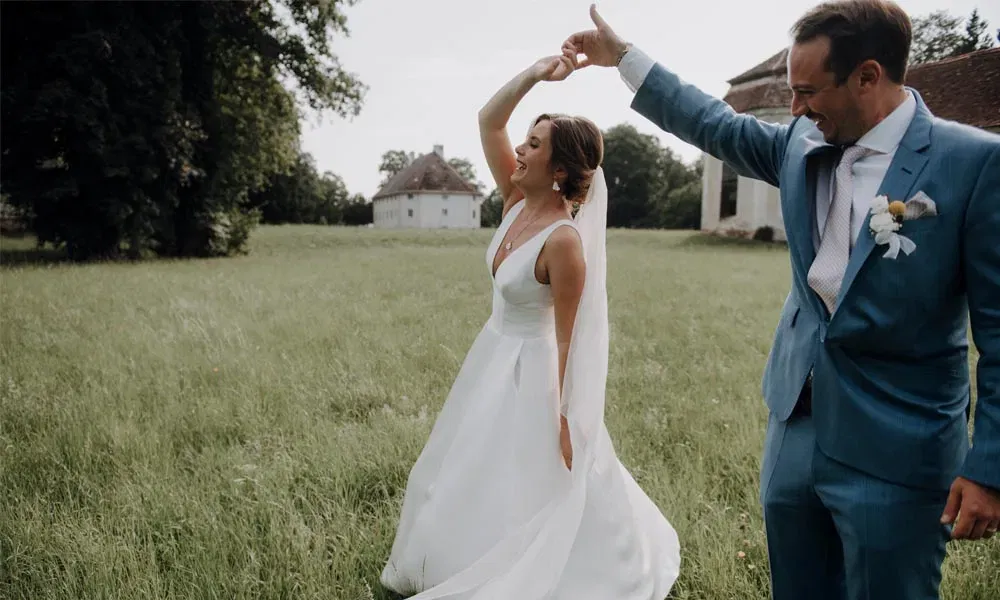 Hochzeitspaar genießt einen fröhlichen Tanzmoment im Freien, umgeben von Natur und alten Gebäuden.