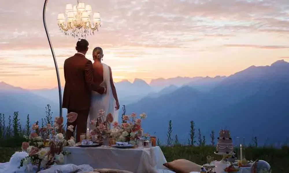 Romantische Szene: Paar in Hochzeitskleidung genießt den Ausblick auf Berge und Sonnenuntergang bei elegant gedecktem Tisch.