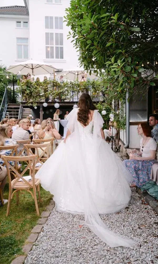 Eine Braut im weißen Prinzessinnenkleid mit langer Schleppe läuft bei der Eröffnungsgala der Villa Lindström durch den Garten, während Gäste applaudieren.