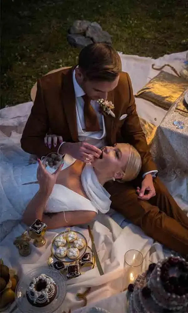 Hochzeitspaar genießt süße Leckereien auf Picknickdecke, eng umschlungen und entspannt.