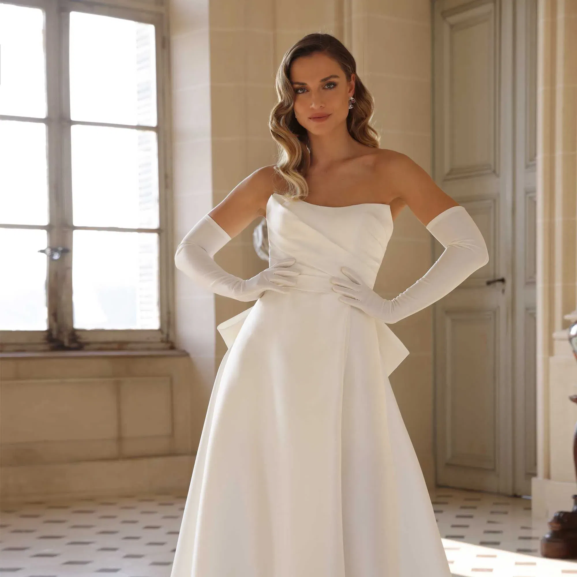 Moderne Brautmode: schlichtes, trägerloses Hochzeitskleid in A-Linie mit eleganten Accessoires und glamourösen Wellen im Haar.