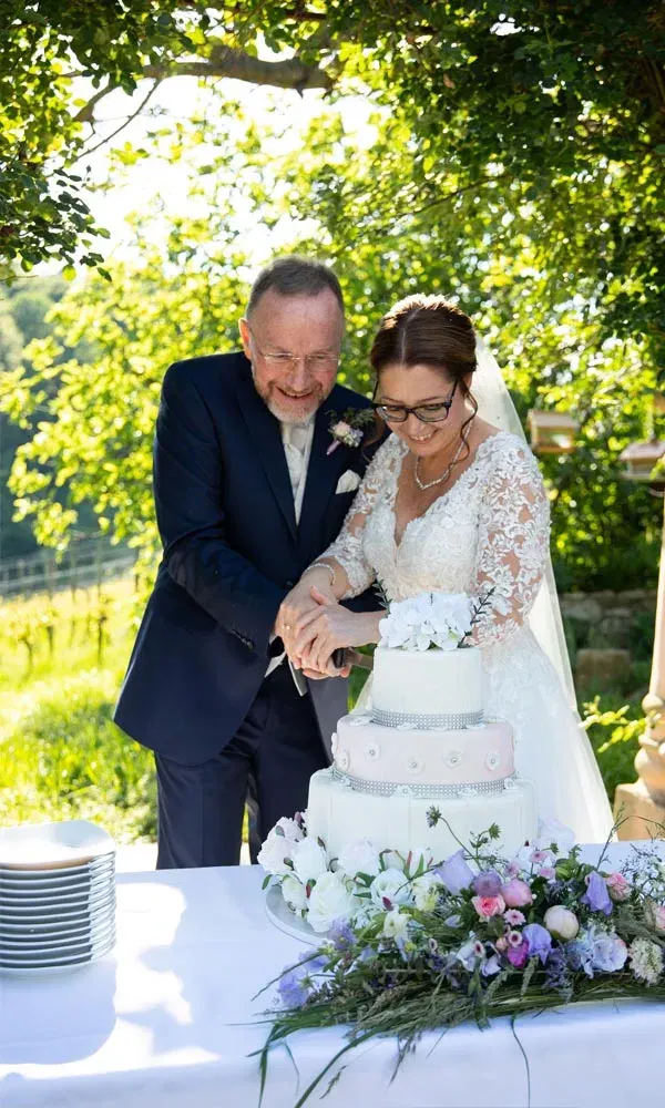 Braut und Bräutigam schneiden gemeinsam lachend ihre Hochzeitstorte mit Blumen-Dekoration im Freien an.