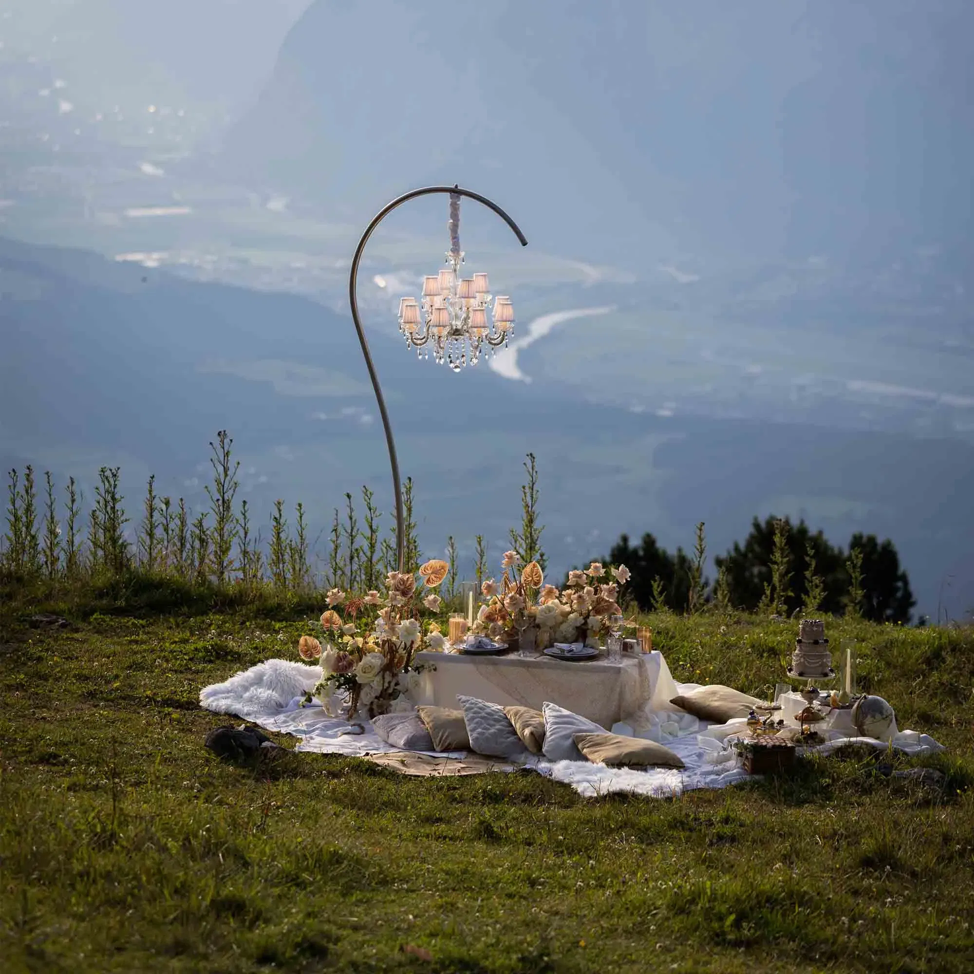 Romantisches Outdoor-Picknick mit Kissen, Blumendeko, Torte und schwebendem Kronleuchter vor Bergkulisse.