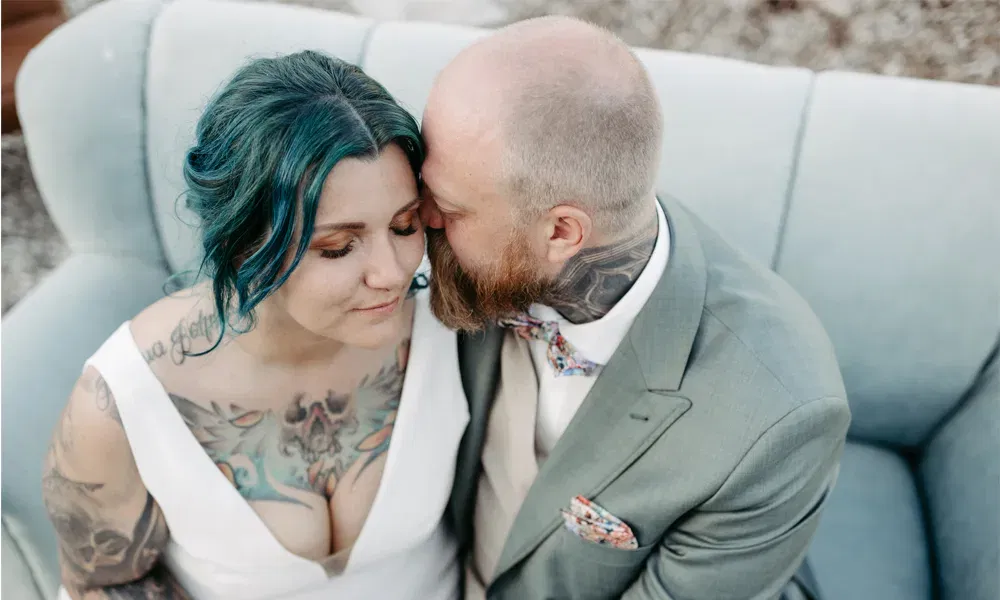 Braut mit türkisfarbenem Haar und Tattoos sitzt neben ihrem Bräutigam, der ihr zärtlich einen Kuss auf die Wange gibt.