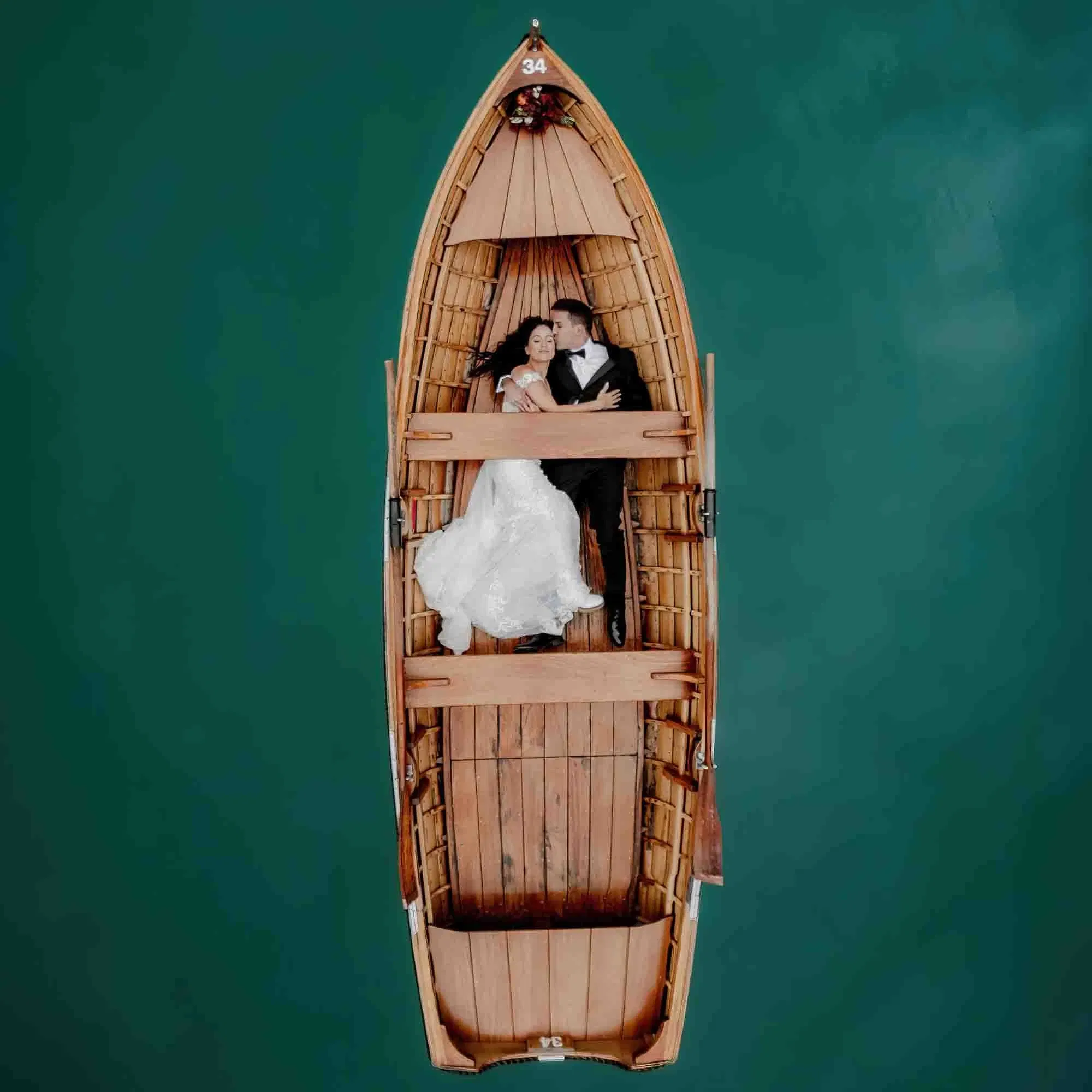 Romantisches Brautpaar liegt eng umschlungen in einem Holzboot auf türkisgrünem Wasser – aufgenommen aus der Vogelperspektive.