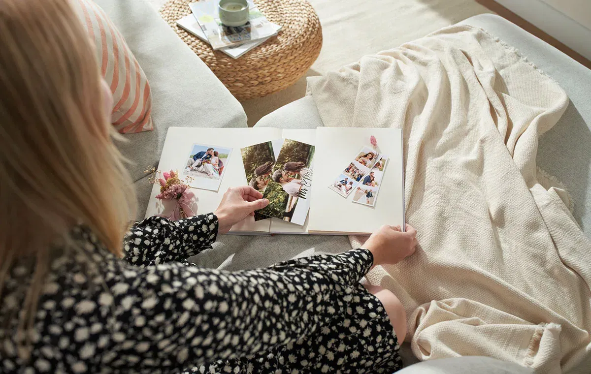 Eine Frau sitzt auf einem Sofa und schaut sich ein liebevoll gestaltetes Hochzeitsalbum mit mehreren Fotos und getrockneten Blumen an.
