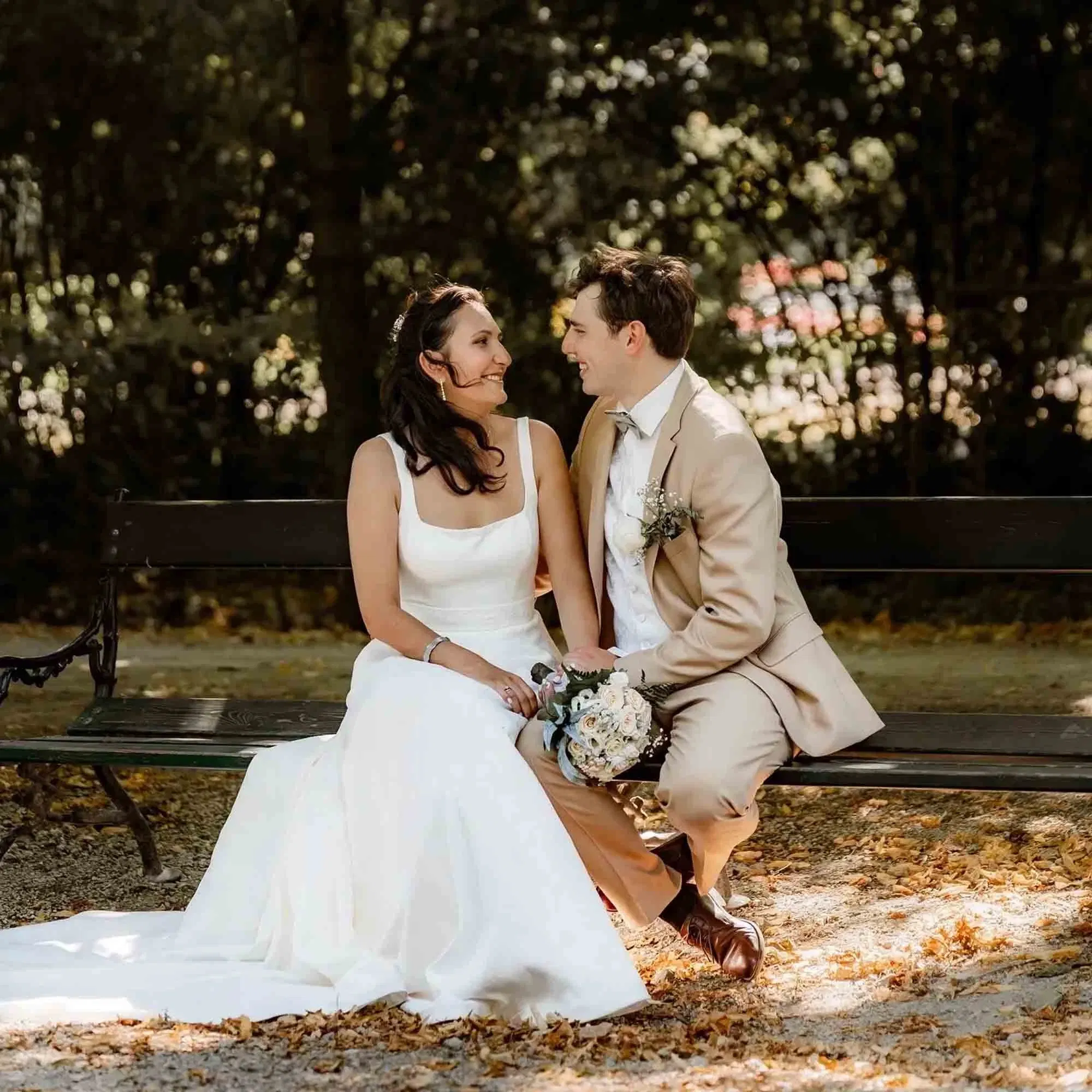 Braut und Bräutigam sitzen verliebt auf einer Parkbank unter Bäumen und blicken sich lächelnd an.
