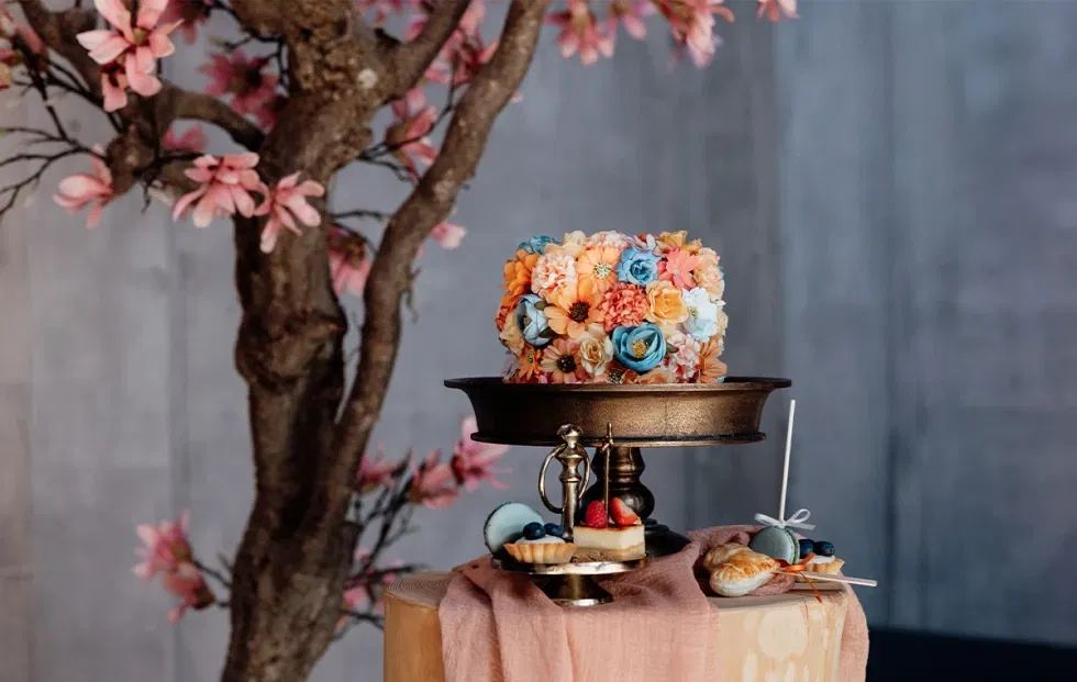 Hochzeitstorte mit bunten Blüten und feinen Gebäckstücken auf einem Desserttisch mit Naturdekoration.