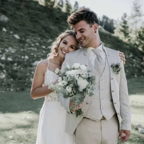 Braut und Bräutigam umarmen sich glücklich im Grünen, sie hält einen weißen Blumenstrauß.