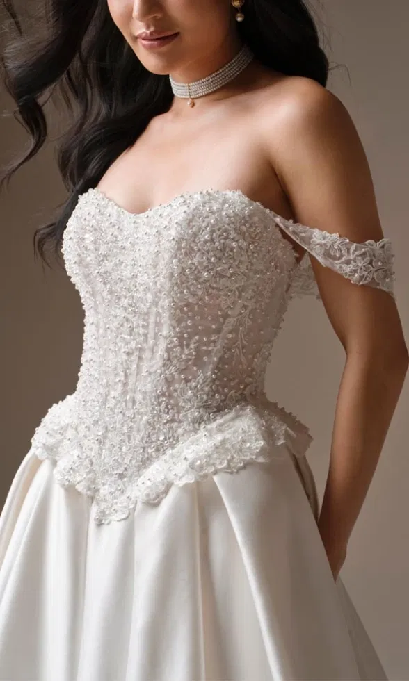 Detailaufnahme eines Brautmodells in einem opulenten Ballkleid mit trägerlosem Korsagenoberteil, dicht besetzt mit Perlen und floraler Spitze, ergänzt durch zarte Spitzenärmel, funkelnden Choker und fließenden Rock vor neutralem Studiohintergrund.