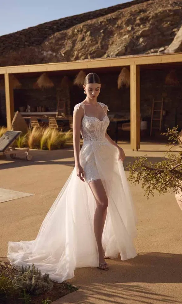 Brautmodell trägt ein modernes Brautkleid mit kurzem Spitzenkleid und abnehmbarem Tüllüberrock, feinen Trägern und fließender Schleppe, inszeniert in einer warmen, naturnahen Outdoor-Location mit minimalistischer Architektur.