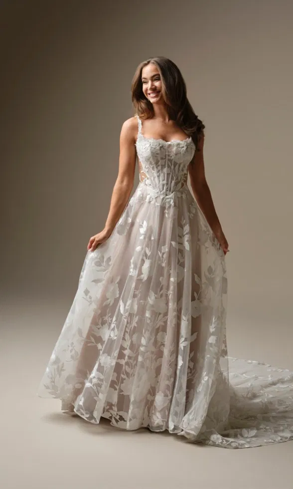 Brautmodell trägt ein elegantes A-Linien-Brautkleid mit feinen Spaghettiträgern, transparentem Korsagenoberteil und floraler Spitze, locker fallender Tüllrock mit Schleppe, inszeniert vor neutralem Studiohintergrund.