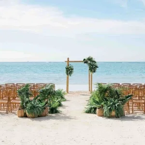 Trauungssetup am Strand mit Holzstühlen, grünem Palmschmuck und einem schlichten Traubogen vor dem Meer.
