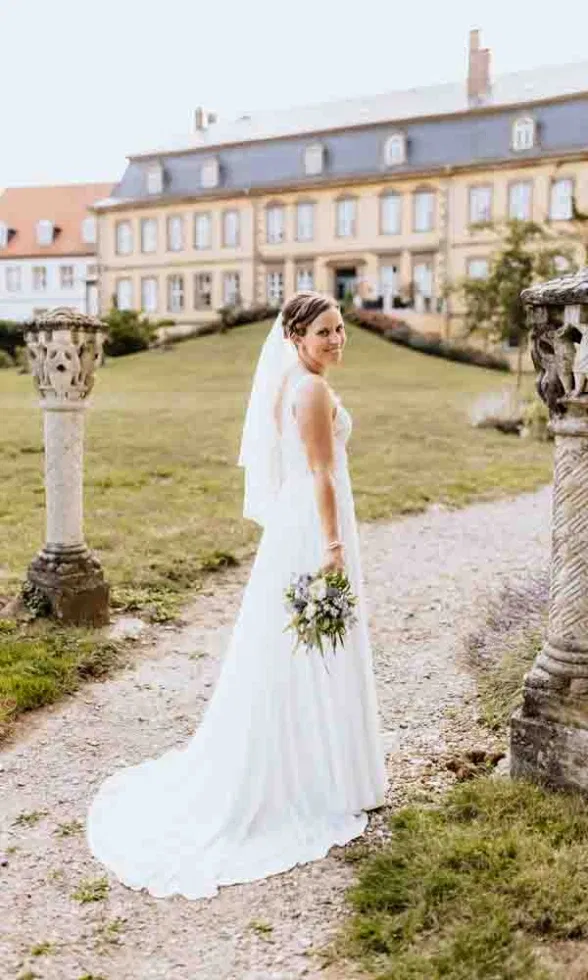 Die Braut dreht sich lächelnd um und präsentiert ihr fließendes Hochzeitskleid vor der historischen Kulisse.