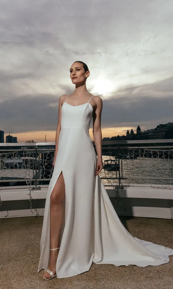 Braut in modernem Spaghettiträgerkleid mit hohem Schlitz auf einer Terrasse bei Sonnenuntergang.