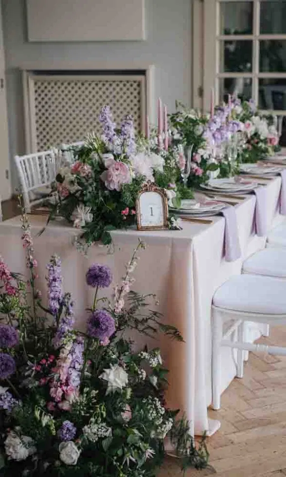 Aufwendig dekorierte Hochzeitstafel mit üppigen Blumenarrangements in Rosa, Weiß und Flieder sowie eleganten Kerzen und einer Tischkarte mit der Nummer 1.