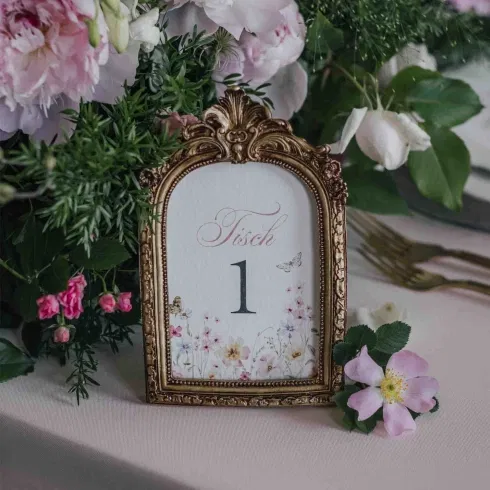 Eleganter Tischaufsteller in goldverziertem Rahmen mit der Aufschrift „Tisch 1“, umgeben von rosa und weißen Blumen in romantischer Hochzeitsdeko.