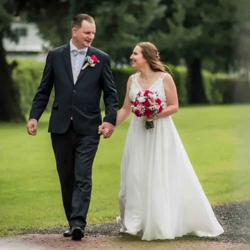 Braut und Bräutigam gehen Hand in Hand über einen Gartenweg und lächeln sich liebevoll an.