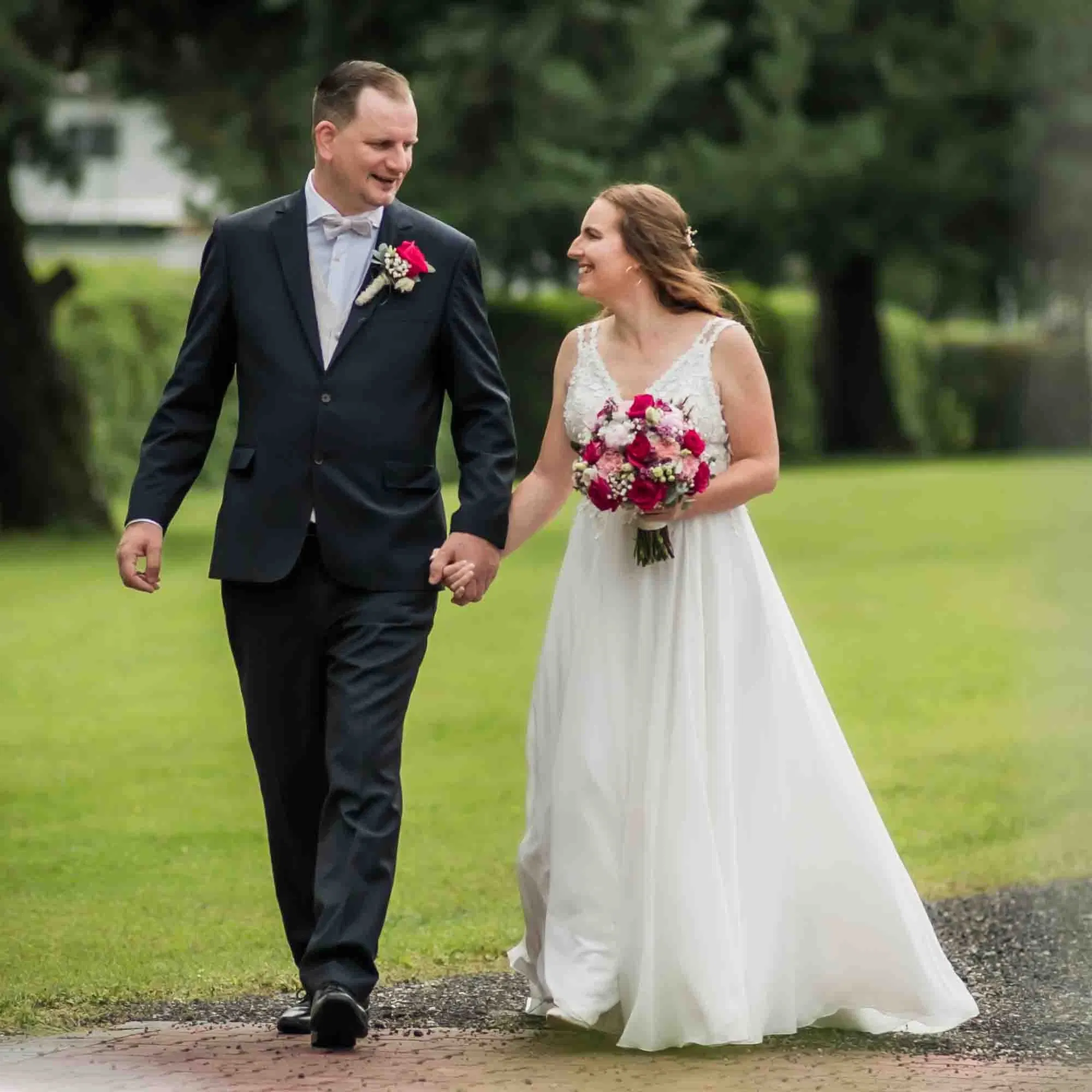 Braut und Bräutigam gehen Hand in Hand über einen Gartenweg und lächeln sich liebevoll an.