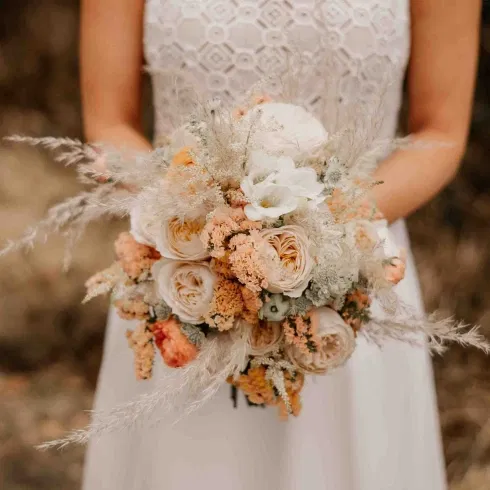 Braut hält einen natürlichen Boho-Brautstrauß aus cremefarbenen Rosen, Pfirsichtönen und zarten Trockenblumen.