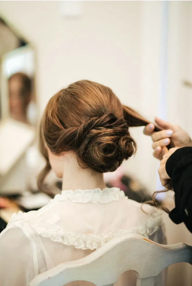 Braut sitzt auf einem Stuhl, während ihre Haare zu einem eleganten, lockeren Dutt gestylt werden.