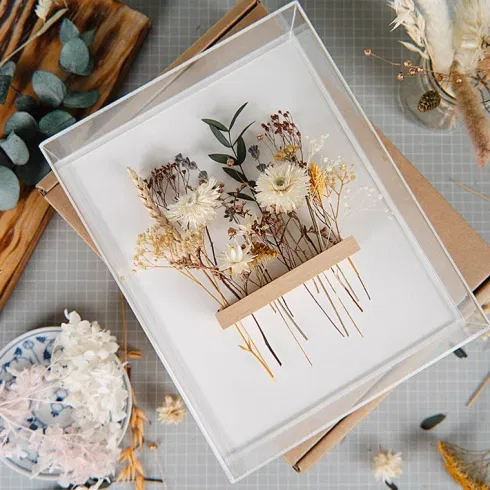 Dekorativer DIY-Bilderrahmen mit zarten getrockneten Blumen in Naturtönen auf weißem Hintergrund, umgeben von Bastelmaterialien und Werkzeugen