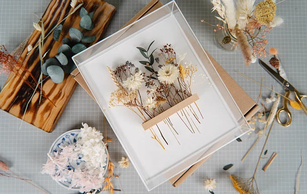 Dekorativer DIY-Bilderrahmen mit zarten getrockneten Blumen in Naturtönen auf weißem Hintergrund, umgeben von Bastelmaterialien und Werkzeugen