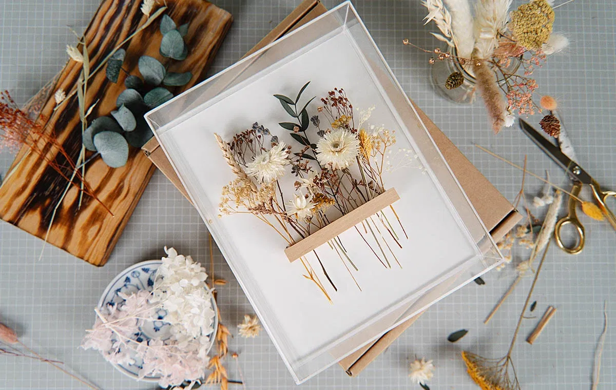 Dekorativer DIY-Bilderrahmen mit zarten getrockneten Blumen in Naturtönen auf weißem Hintergrund, umgeben von Bastelmaterialien und Werkzeugen