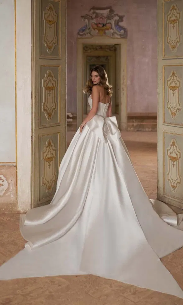 Rückenansicht eines Brautmodells in einem eleganten Brautkleid mit Korsagenoberteil, voluminösem Rock, langer Schleppe und markanter Schleife am Rücken, inszeniert in einem historischen Saal mit aufwendig verzierten Türen und pastellfarbenen Wandmalereien.