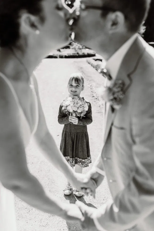 Schwarz-Weiß-Foto eines Brautpaars, das sich küsst, während ein lächelndes Kind mit Blumenstrauß zwischen ihnen steht.