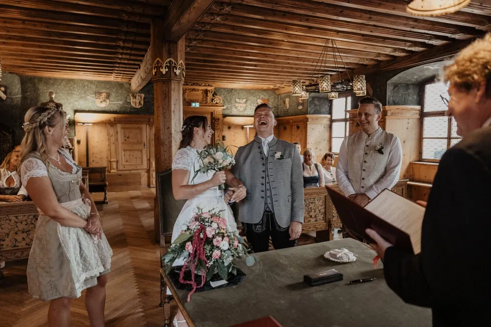 Fröhliches Brautpaar in Tracht lacht während der standesamtlichen Trauung in einem holzgetäfelten Raum.