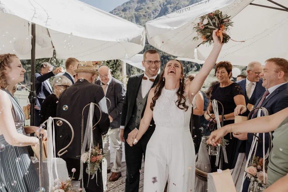 Frisch vermähltes Paar geht lachend durch ein Spalier aus Gästen; die Braut hebt jubelnd ihren Brautstrauß in die Luft, während Seifenblasen und Blütenkonfetti durch die Sonne schweben.