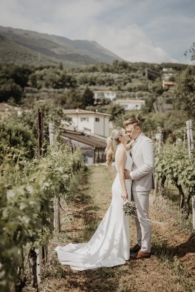 Ein Brautpaar küsst sich in einem sonnigen Weinberg mit Blick auf die Hügel und Häuser im Hintergrund.