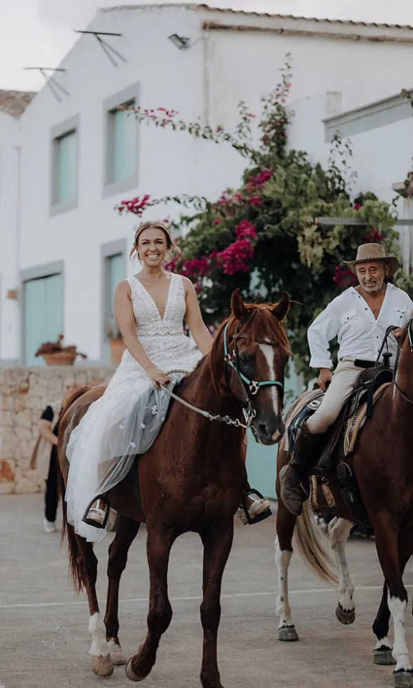 Die Braut reitet auf einem braunen Pferd durch einen Hof mit weißen Häusern und Bougainvillea, während ein Reiter neben ihr herkommt.