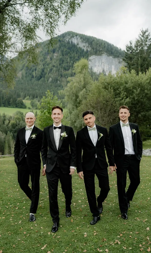 Bräutigam und Trauzeugen im schwarzen Smoking gehen gemeinsam über eine Wiese vor alpiner Bergkulisse.