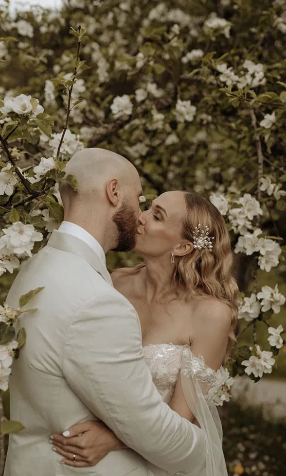 Braut und Bräutigam küssen sich unter einem blühenden Baum, umgeben von weißen Frühlingsblüten.