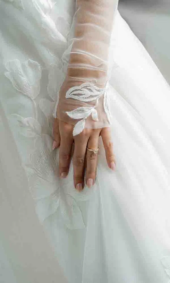 Detailaufnahme der Brauthand mit zartem Spitzenhandschuh und Ring, ruhend auf einem floralen Brautkleid aus Tüll.