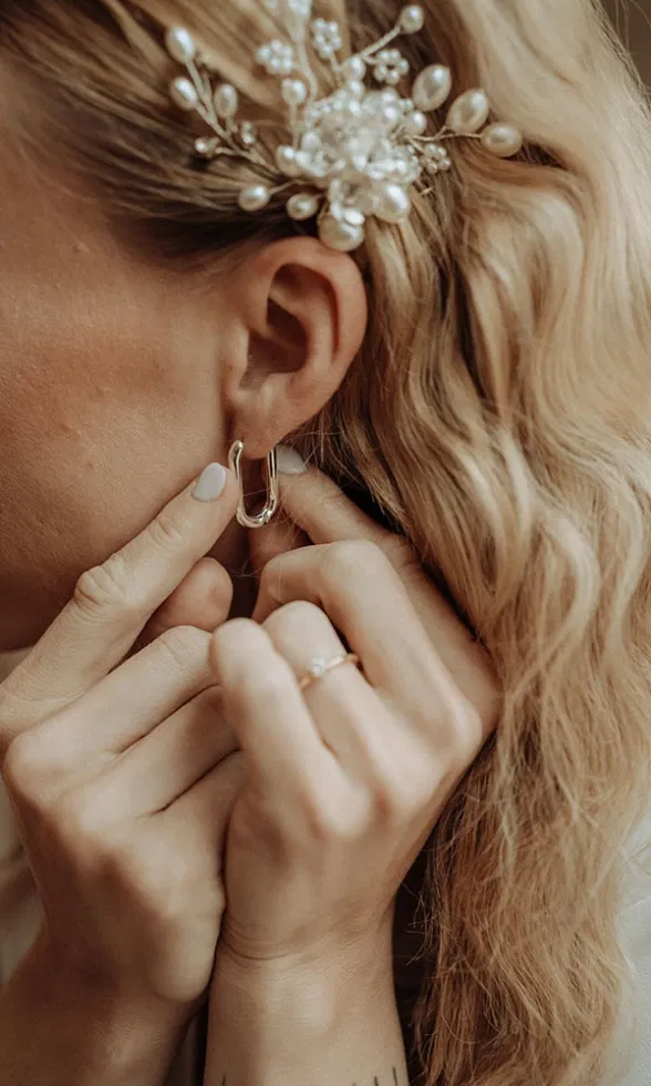 Detailfoto des Gesichtsbereichs: Die Braut befestigt ihren Ohrring, während ein filigraner Perlenschmuck ihr Haar ziert.