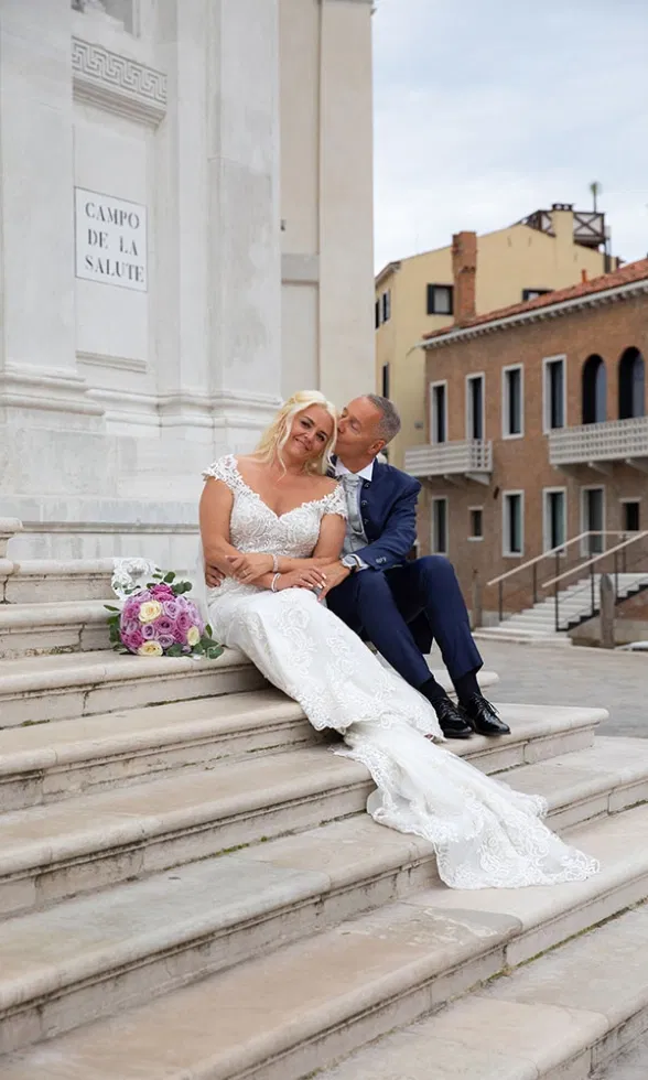 Braut und Bräutigam sitzen entspannt auf den Stufen vor einer historischen Kirche, der Bräutigam gibt der Braut einen liebevollen Kuss auf die Wange, während ihr Spitzenbrautkleid mit Schleppe elegant über die Stufen fällt und ein pastellfarbener Brautstrauß neben ihnen liegt.