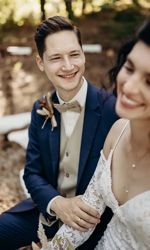 Brautpaar sitzt nah beieinander im Wald, der Bräutigam lächelt die Braut liebevoll an, intime und natürliche Hochzeitsaufnahme.