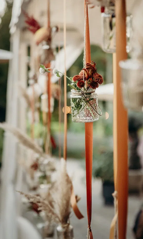 Hängende Hochzeitsdekoration mit kleinen Glasvasen, herbstlichen Blumen und Bändern in warmen Rost- und Naturtönen.