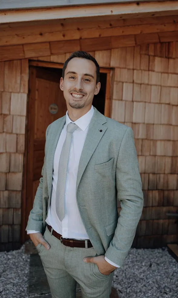 Der Bräutigam lächelt in seinem hellen Anzug in die Kamera und steht entspannt vor einer Holzhütte.