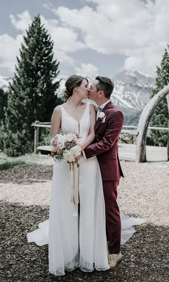 Braut und Bräutigam küssen sich zärtlich vor alpiner Kulisse, die Braut hält einen Blumenstrauß, im Hintergrund Berge und Tannen.